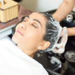 Face of relaxed young Latin American woman enjoying hair washing in salon. Hairdresser washing hair of woman with shampoo